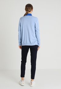 Jasnoniebieski sweter z dzianiny o prostym kroju, z prążkowanym dołem, noszony na ciemnoniebieską koszulę z kołnierzykiem, w połączeniu z czarnymi spodniami i białymi butami.