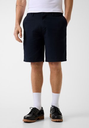 Uomo che indossa pantaloncini neri fino al ginocchio, calzini bianchi, scarpe nere con lacci e canottiera bianca, in piedi con le mani nelle tasche.