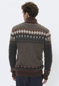 Hombre de espaldas vistiendo un suéter acogedor marrón con un patrón de rombos y puntos, y pantalones oscuros, sobre un fondo blanco.