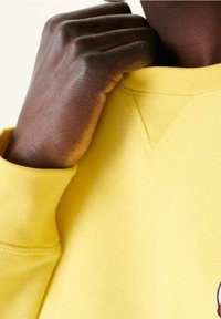 Main à la peau foncée ajustant le col d'un sweat-shirt jaune avec des détails de coutures visibles et des poignets côtelés.