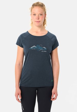 Femme aux cheveux blonds bouclés portant un t-shirt bleu marine avec un motif de montagne et un pantalon noir, sur fond uni.