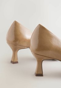 Beige korkokenkiä kiiltävällä pinnalla ja kulmikkailla korkoilla, aseteltuna pehmeää, neutraalia taustaa vasten.