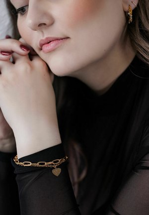 Gold chain bracelet with a heart charm, worn on a wrist beside a black long-sleeved top. Smooth texture and interconnected links visible.