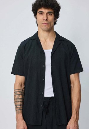 Homme aux cheveux foncés et bouclés porte une chemise noire à manches courtes boutonnée sur un débardeur blanc, mettant en avant un tatouage géométrique sur son avant-bras gauche.