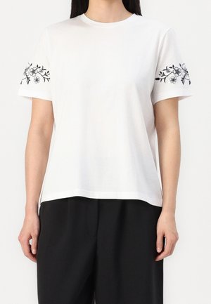 Hvid t-shirt i blød bomuld med sort blomsterbroderi ved ærmerne. Klassisk rund halsudskæring og afslappet pasform. Sammen med sorte bukser.