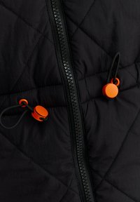 Nahaufnahme von schwarzem gestepptem Stoff mit einem zentralen schwarzen Reißverschluss und zwei schwarzen Kordeln mit leuchtend orangefarbenen Kunststoffstoppern.