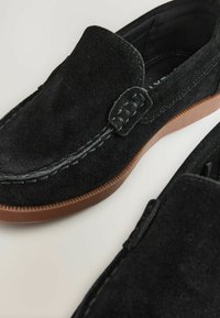 Mocassins en daim noir avec une texture lisse, des détails cousus et des semelles en caoutchouc marron, conçus pour un enfilage facile et un confort optimal.