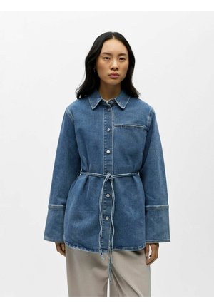 Kobieta o długich czarnych włosach, ubrana w niebieską, jeansową kurtkę zapinaną na guziki z paskiem przewiązanym w talii oraz jasne spodnie, na jednolitym tle.