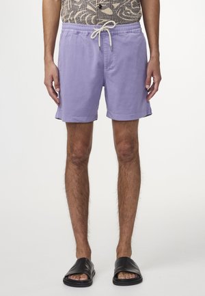 Lila Baumwollshorts mit elastischem Bund und Kordelzug. Sie verfügen über seitliche Taschen und eine legere Passform. Getragen mit schwarzen Rutsch-Sandalen.