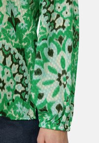 Grüne Bluse mit floralen Motiven aus einem leichten, strukturierten Stoff. Sie zeichnet sich durch eine Mischung aus dunkelgrünen und hellblauen Mustern, gerafften Ärmeln und einem lockeren Schnitt aus.