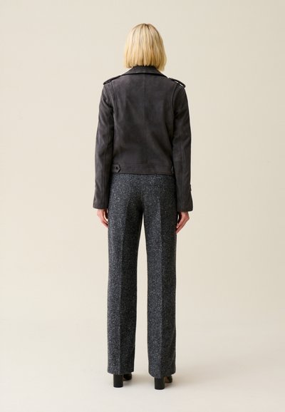Giubbotto di pelle nera con una vestibilità aderente, spalline dettagliate sulle spalle, abbinato a pantaloni grigi a gamba larga e texture. Sfondo neutro.