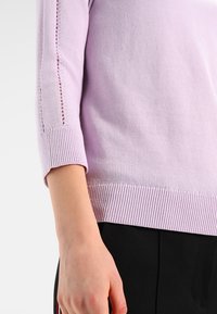 Jasnofioletowy dzianinowy top z rękawami do łokcia, z prążkowanym dołem i subtelnymi koronkowymi zdobieniami po bokach.