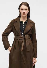 Femme portant un trench-coat en suède marron foncé ceinturé par-dessus une chemise noire boutonnée, debout contre un fond clair uni.