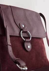 Handtas van bordeauxrood leer en suède met een gestructureerde afwerking, voorzien van een zilveren, cirkelvormig metalen detail en verstelbare band.
