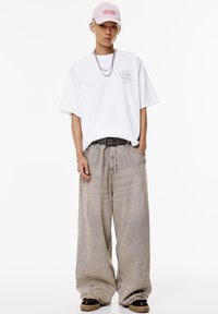 T-shirt bianca oversize con grafica, cappellino rosa chiaro, pantaloni in denim beige larghi, cintura nera con borchie e calzature con suola scura.