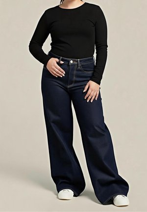 Vrouw draagt een zwarte top met lange mouwen, hoge taille wijde donkeblauwe jeans en witte sneakers, staand tegen een beige achtergrond.