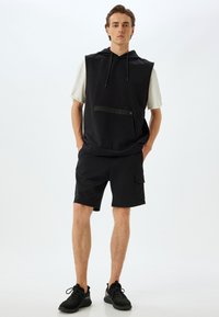 Sudadera sin mangas negra con un bolsillo delantero y cordones, combinada con pantalones cortos negros con bolsillos laterales. Llevado con zapatillas deportivas negras.