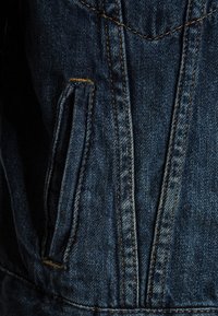 Sekce džínové bundy s tmavomodrou látkou a oranžovými švy a přední kapsou. Výrazná texturovaná vazba a viditelné švy.