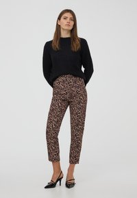 Donna in piedi con le mani dietro la schiena, che indossa un maglione nero a trama, pantaloni a tre quarti con stampa leopardata e scarpe con tacco nere.