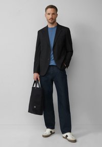 Czarny żakiet na niebieskiej koszulce, zestawiony z ciemnoniebieskimi dżinsami i białymi trampkami z brązowymi akcentami. Trzyma czarną torbę na ramię.