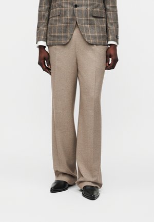 Homme portant un pantalon beige à jambes larges, des chaussures noires pointues, et un blazer marron à carreaux avec des poignets blancs, se tenant devant un fond uni.