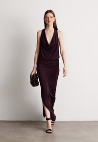 Μακριά, εφαρμοστή, χωρίς μανίκια φόρμα σε σκούρο μωβ ύφασμα με ριγμένο μπούστο; συνδυασμένη με μια μαύρη τσάντα και μαύρες σαγιονάρες με λουριά.