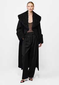 Długi czarny płaszcz z teksturowanym czarnym futrzanym kołnierzem i mankietami, noszony na przezroczystej czarnej bluzce, w połączeniu z czarnymi spodniami z wysokim stanem i sandałami na obcasie.