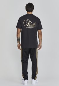 Camiseta de algodón negra con logo impreso en dorado en la parte posterior. Combinada con pantalones de chándal negros, acentuados por rayas doradas en los lados y zapatillas blancas.