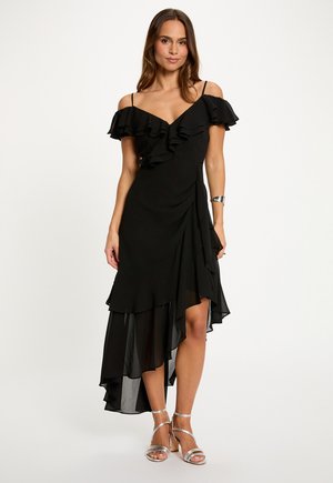 Donna con un vestito nero con spalle scoperte e volant, orlo asimmetrico e sandali argentati con tacco alto, in piedi davanti a uno sfondo semplice.