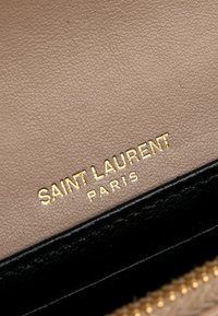 Cuir beige texturé avec logo doré "Saint Laurent Paris" au-dessus du bord en cuir noir et dents de fermeture éclair dorées.