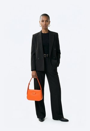 Vrouw draagt een zwarte blazer, zwart topje, zwart wijde pantalon met krijtstrepen, zwarte schoenen, gouden oorringen en houdt een feloranje handtas vast.