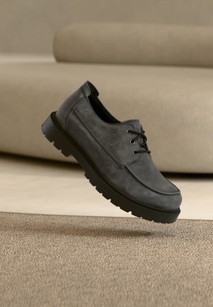 Grauer Veloursleder-Schnürschuh mit dicker schwarzer Sohle, der über einem strukturierten beigen Teppich in einem neutral gehaltenen Raum schwebt.