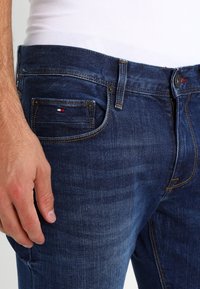 Jeans de mezclilla azul oscuro con corte recto, costuras en contraste y una pequeña etiqueta con el logo en el bolsillo trasero. Cierre con cremallera y botón.