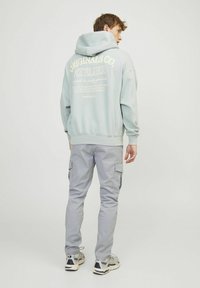 Helles blaues Kapuzenshirt mit bedrucktem Text auf der Rückseite, kombiniert mit hellgrauen Cargo-Hosen. Das Outfit beinhaltet Sneaker in einer neutralen Farbpalette.