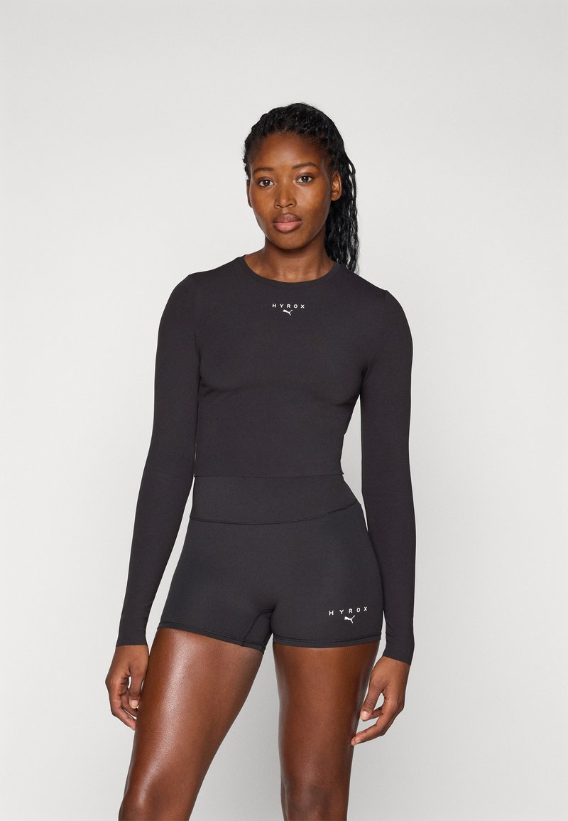 Ensemble composé d'un haut à manches longues noir et d'un short, réalisé en tissu extensible, avec une coupe ajustée et des accents de logo blanc sur le haut et le short.