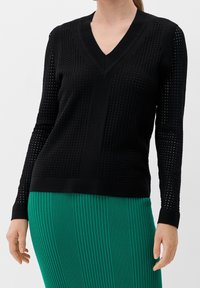 Schwarzer Strickpullover mit V-Ausschnitt, der ein strukturiertes Muster mit Löchern entlang der Ärmel und des Körpers aufweist. Wird über einem grünen plissierten Rock getragen.