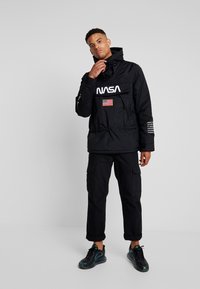 Zwarte gewatteerde jas met een capuchon, voorzien van een opvallend wit "NASA" logo, een patch van de Amerikaanse vlag en een elastische koord met trekker in de taille.