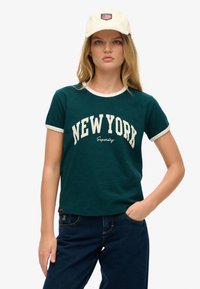 Camiseta de algodón verde con cuello blanco, que presenta "NUEVA YORK" en letras color crema. Combinada con jeans de mezclilla azul oscuro.