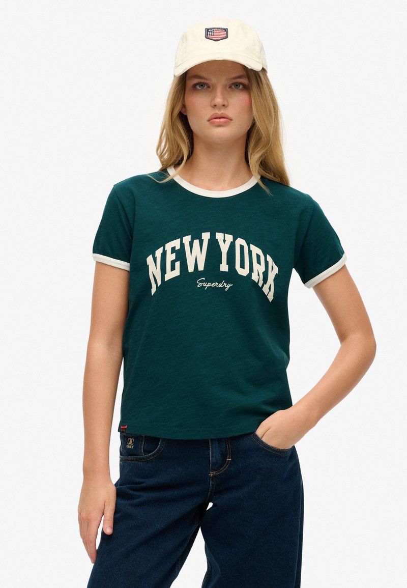 Camiseta de algodón verde con cuello blanco, que presenta "NUEVA YORK" en letras color crema. Combinada con jeans de mezclilla azul oscuro.