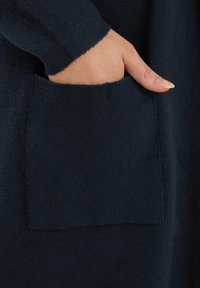Indumento in lana blu navy con una texture morbida, caratterizzato da una grande tasca sul davanti. Primo piano di una mano posizionata nella zona della tasca.
