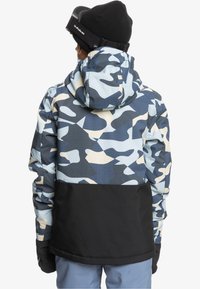 Kamouflagemönstrad jacka i blått, beige och svart med luva. Har en texturerad tygkonstruktion och en kontrasterande svart nederdel.