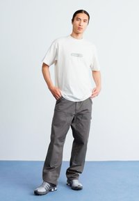 Helebeige puuvillane T-särk, millel on roheline graafika rinnal, koos tumehallide cargo-pükste ja hallide spordijalatsitega.