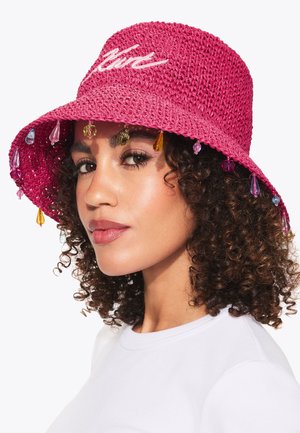 Vrouw met krullend haar draagt een roze gehaakte bucket hat versierd met kleurrijke hangende kralen en een wit shirt.