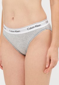 Primo piano di una persona che indossa slip grigi Calvin Klein con una fascia in vita bianca, con il nome del marchio in testo nero.