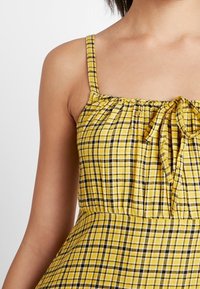 Vestido a cuadros amarillos y negros con tirantes finos y un cuerpo fruncido que presenta un detalle de lazo en la parte delantera. El material parece ligero y texturizado.