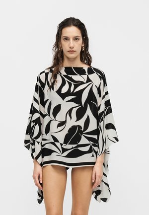 Kvinde iført en løstsiddende sort og hvid bladmønstret top med vide ærmer, stående mod en ensfarvet lys baggrund.