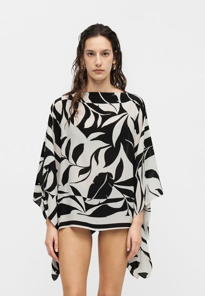 Femme portant un haut ample à motif de feuilles noir et blanc avec des manches larges, debout devant un fond clair uni.