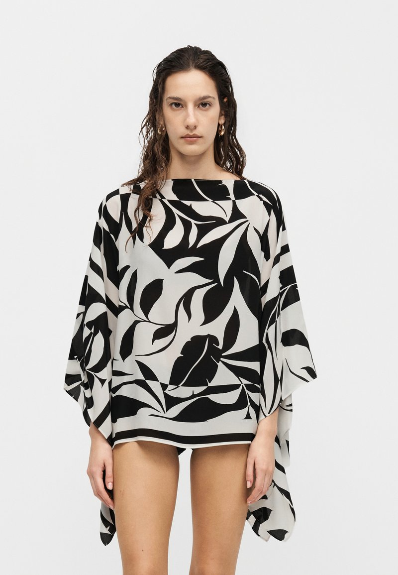 Γυναίκα που φοράει μπλούζα σε άνετη γραμμή με ασπρόμαυρο σχέδιο φύλλων και φαρδιά μανίκια, στέκεται μπροστά σε απλό φωτεινό φόντο.