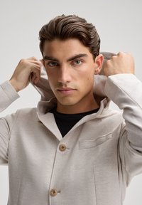 Giacca in misto cotone beige chiaro con cappuccio, dotata di chiusura a doppio bottone e di una piccola tasca sul petto. Texture liscia.