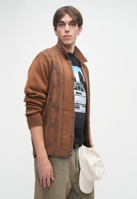 Καφέ πλεκτό κασκόλ με σκουρότερη καφέ σχέδιο, κλείσιμο με κουμπί και φαρδύ γιακά. Φορεμένο πάνω από ένα γραφικό μαύρο t-shirt και συνδυασμένο με μπεζ καπέλο.